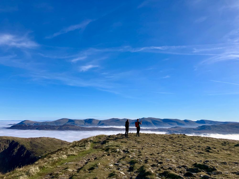 A silhouette of 2 people against the Helvellyn range with blue sky above and cloud below 
