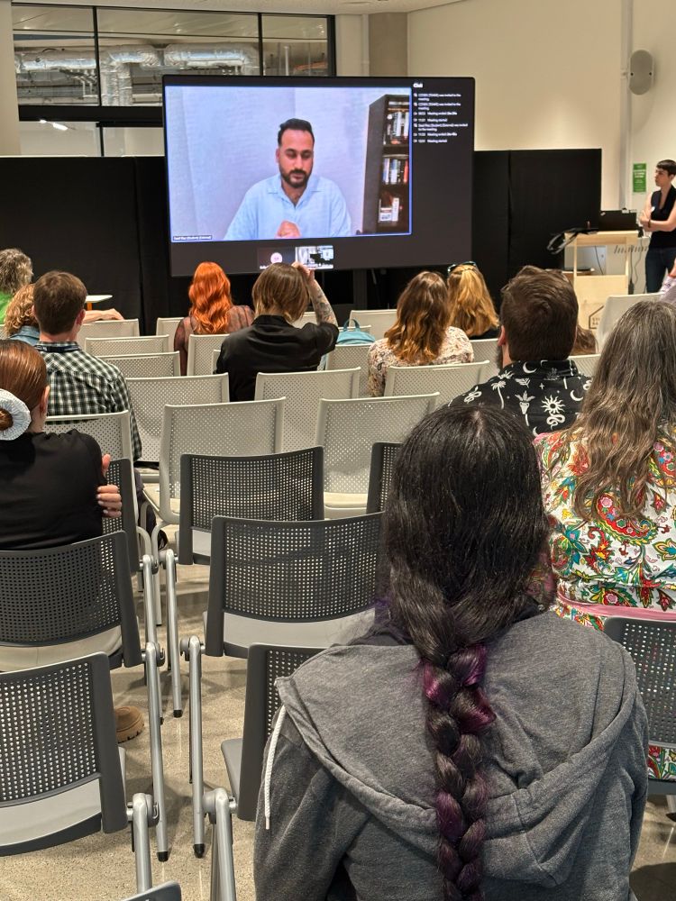 Saad Riaz on screen presenting his work remotely. The phot shows a screen at the front of the room with an audience looking at him in it. He was answering questions about his brilliant work on online discourse in the Noor Mukadam case