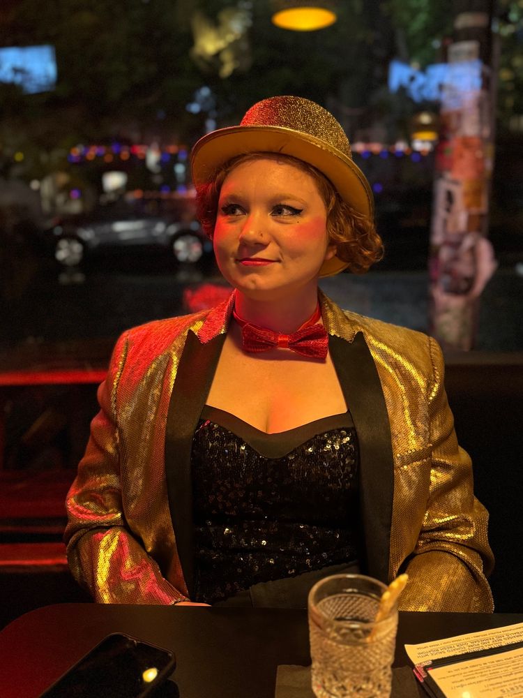 A portrait of a woman dressed up as Columbia from Rocky Horror Picture Show sitting at a table with a cup in front of her