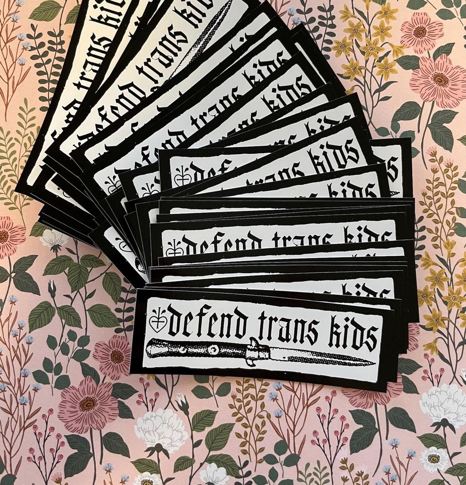 A pile of vinyl stickers fanned out over a floral wall paper print of soft flowers over a pale pink background. The sticker has a black frame around the edge. In the center is a classic switch blade knife. Above is lettering that says “defend trans kids” in an olde English or gothic type face. 