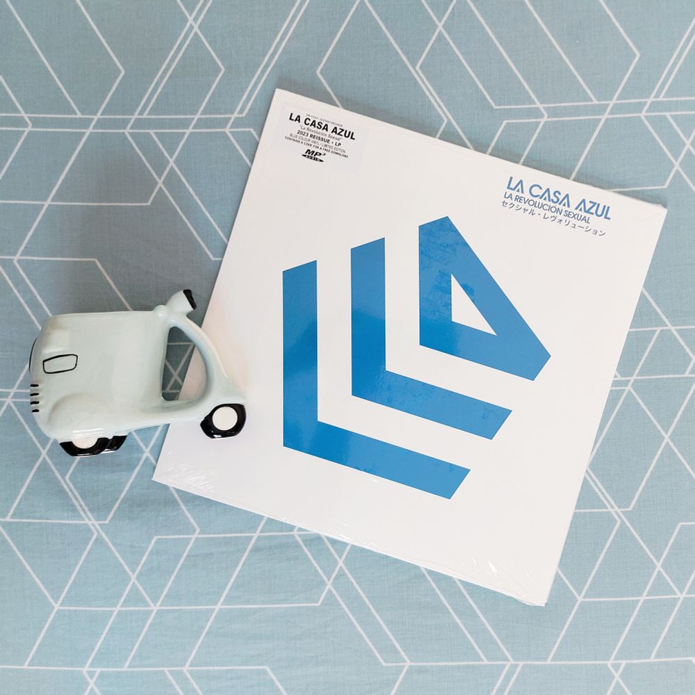 Álbum 'La Revolución Sexual' de La Casa Azul en su versión de vinilo azul de 2023 sobre un fondo también azul con líneas blancas que hacen dibujos geométricos. A la izquierda hay una taza de cerámica con forma de Vespa de color verde claro.