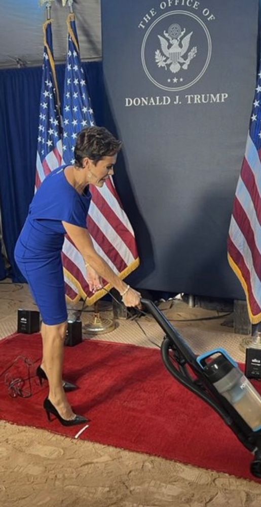 Kari Lake (trump’s maid?) vacuums a red rug placed on sand, while wearing very high heeled shoes 👠