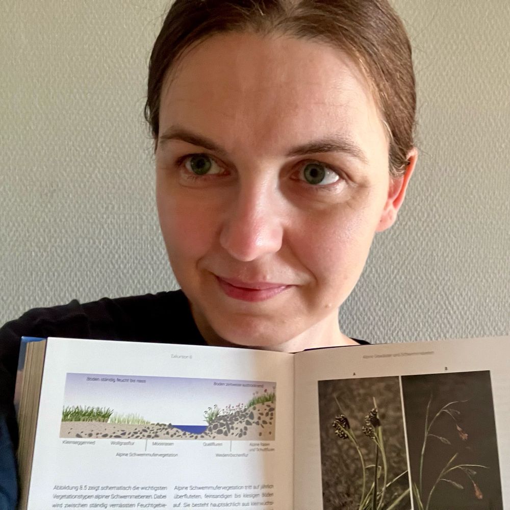 A photo of Dorota Paczesniak (me) holding an open book. On one page there is an illustration of a cross section of an alpine stream and surrounding vegetation. On the other page some photos of alpine plants