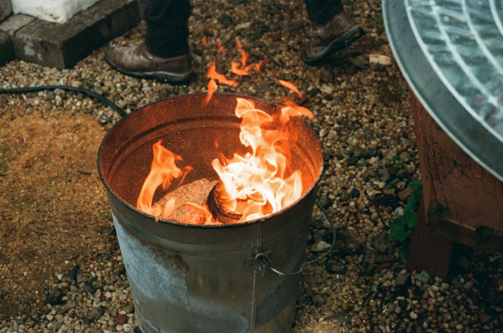 A photograph of a step in the raku pottery firing process. The ground is wet and covered in rocks with a pile of sawdust on the left. In the upper left corner is a hint of the raku kiln, but all thats showing is a few grey bricks. There is a person standing next to the bricks wearing black skinny pants and brown leather work boots. There is a metal bucket in the center containing a big layer of sawdust and a few pottery pieces on fire. They’re red hot and covered in carbon. On the right is a hint of the bottom of a brown wood bench seat and the metal top to the bucket peeking into frame. A small plant growing at the foot of the bench seat adds a small and subdued pop of green.