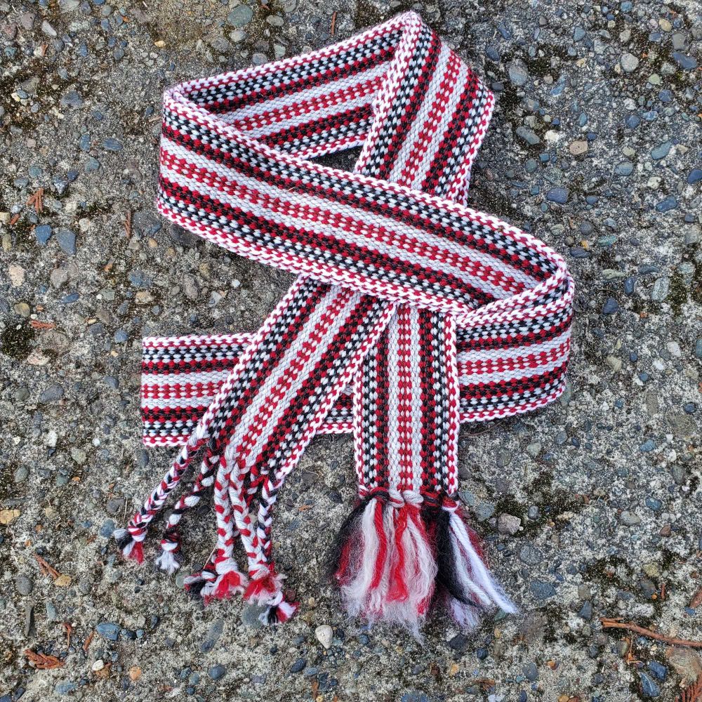 A woven belt folded over it's self. It is red, white, gray, and black. The edges show a kind of gray, black and then red and black pattern. The center is pale gray with a red cross like center pattern. 