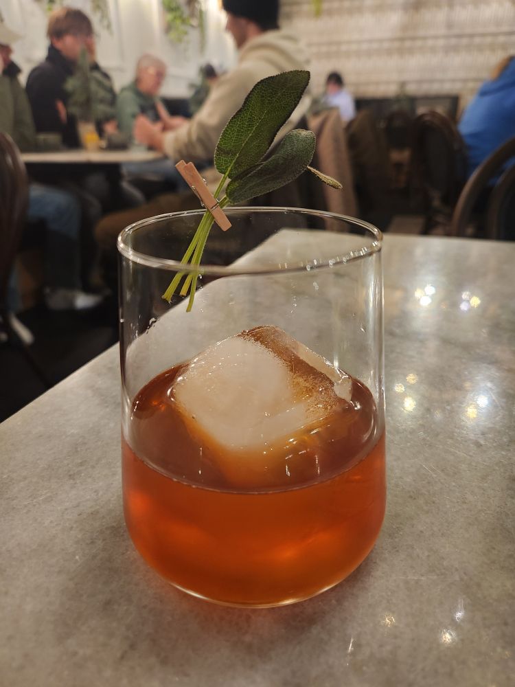 A gorgeous deep orange cocktail with a giant ice cube and a sprig of sage on the rim
