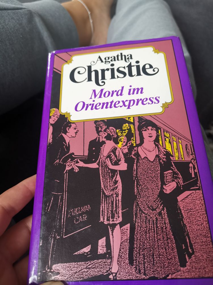 Mord im Orientexpress von Agatha Christie
Das Buchcover ist lila 💜
Man sieht Menschen am Bahnsteig
Frauen in schicken Kleidern, Männer in Anzügen.
Sie flanieren, unterhalten sich, der  Schaffner steht dabei und eine Frau schaut aus dem Zugfenster. 