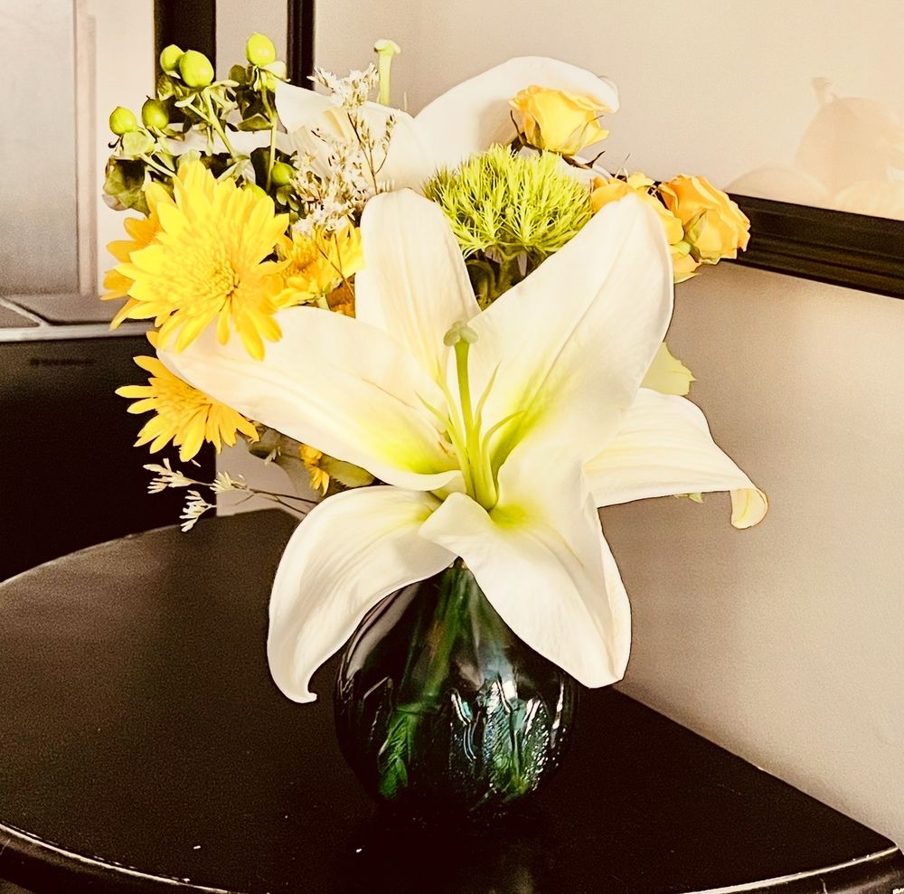Yellow daisies, white lilies, yellow roses, green hypericum, and green ball dianthus in a teal Agavero Tequila liqueur bottle on a small black half table. 