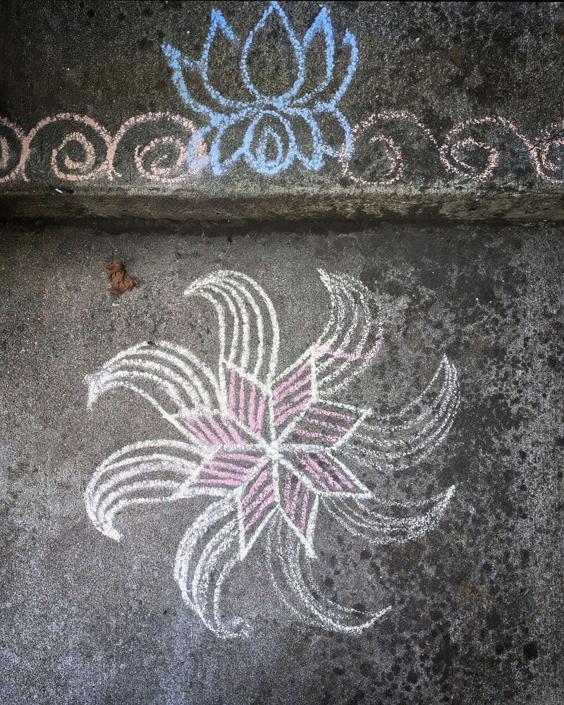 A chalk rangoli design (Indian folk art) on a concrete sidewalk and step. The lower design is white with pink in the center. The upper design is blue in the center and light orange on the sides.