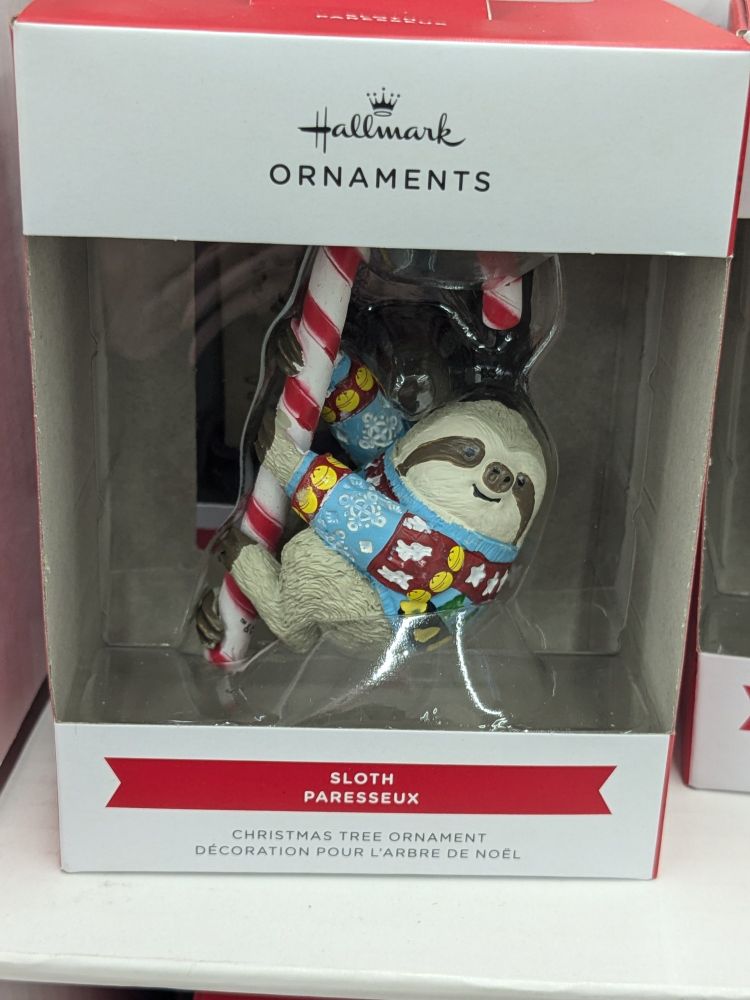 Picture of a hallmark ornament depicting a three-toed sloth holding on to a candy cane and wearing a blue, red, and yellow Christmas sweater.