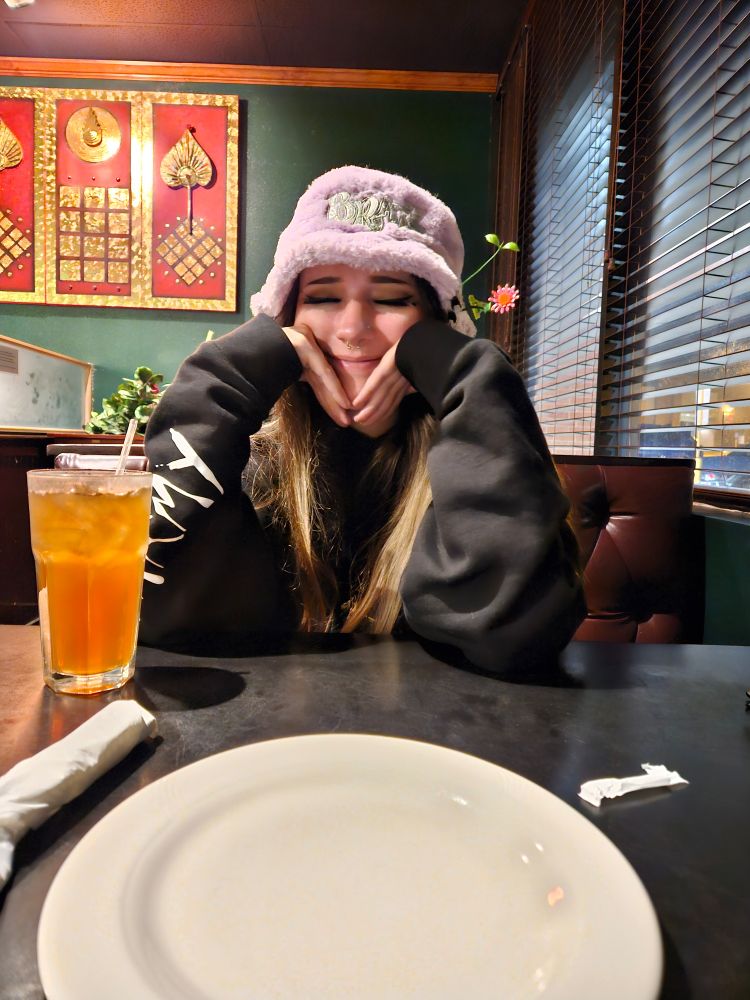 My girlfriend, Anne sitting at a thai restaurant table with her elbows resting on the surface and her hands squishing her cheeks while she smiles sweetly. She has long hair and is wearing a lavender bucket hat and a black Cyclops Army sweatshirt. In front of her is a glass of iced tea and an empty white plate

