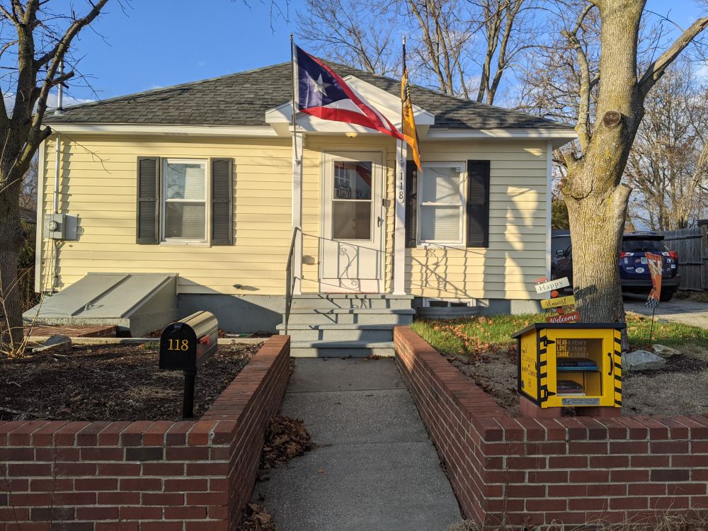 A yellow house with black shutters with a small little free library in the right corner of the yard.