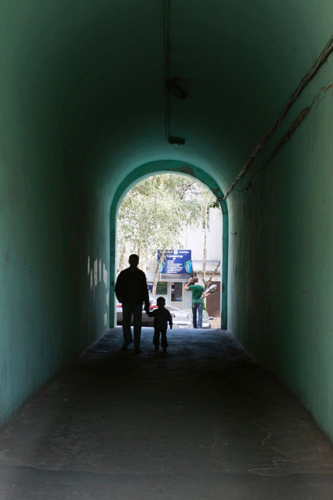 Kyiv 2010. Father and child walking hand in hand out from an opening in a city block pit into the street. A man with flowers in his hand out in the sun 