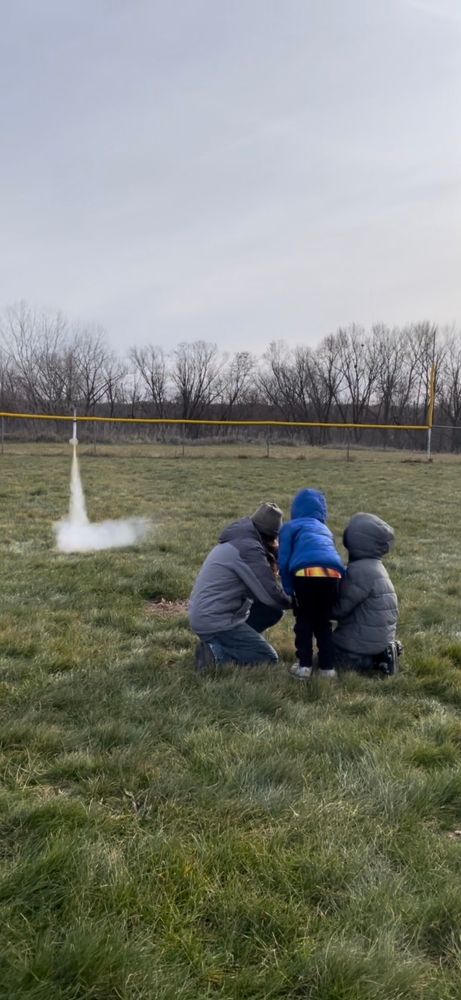 Three guys watching a model rocket during liftoff phase, one person is middle age and the other two are under 12
