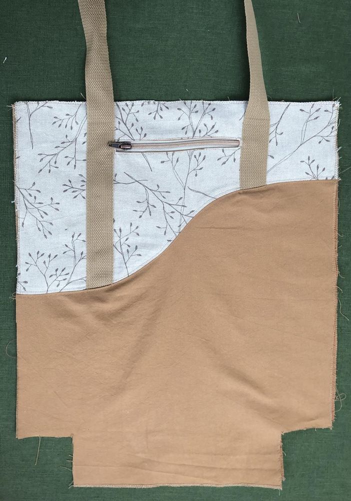 Halbe Einkaufstasche aus Stoff liegt auf grünem Tuch. Die Taschenhälfte ist durch eine bogenförmige Linie getrennt, in der oberen Hälfte ist sie beige und ocker, Gurtband ist sichtbar. Unterer Teil ist ocker Stoff, das Gurtband ist im unteren Teil nicht sichtbar. 