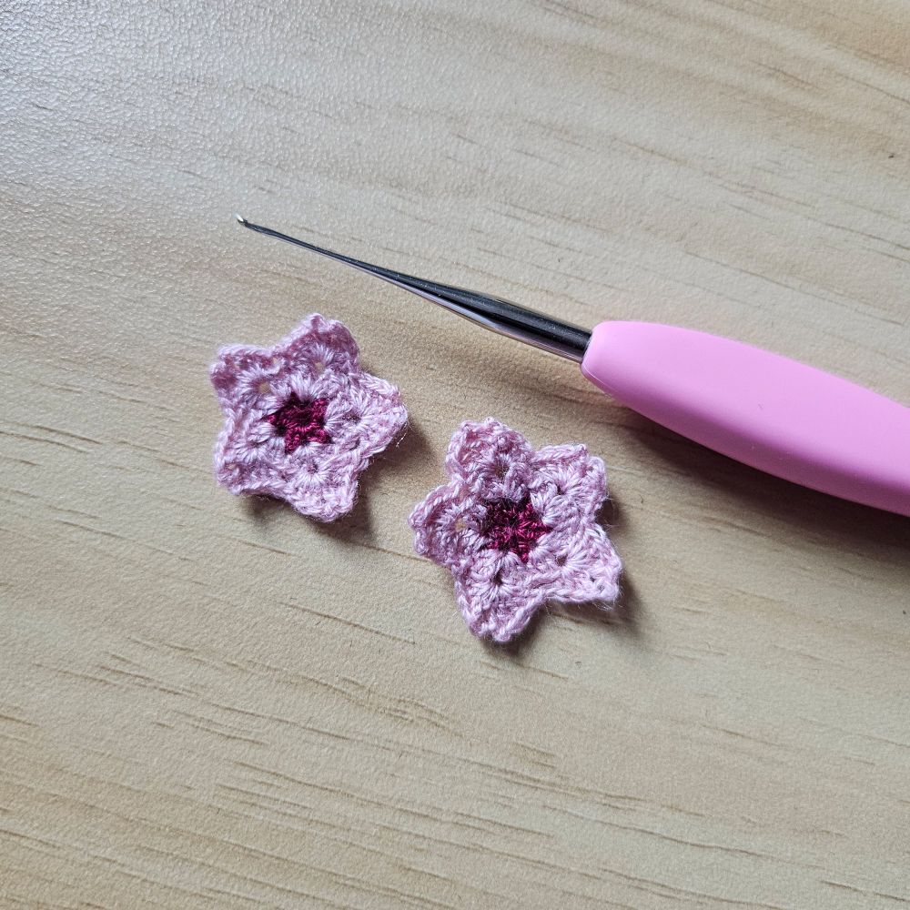 two micro crochet flowers on a surface with a wood grain pattern, next to a tiny crochet hook. the flowers are pink with dark red centres and notched petals, like cherry blossoms