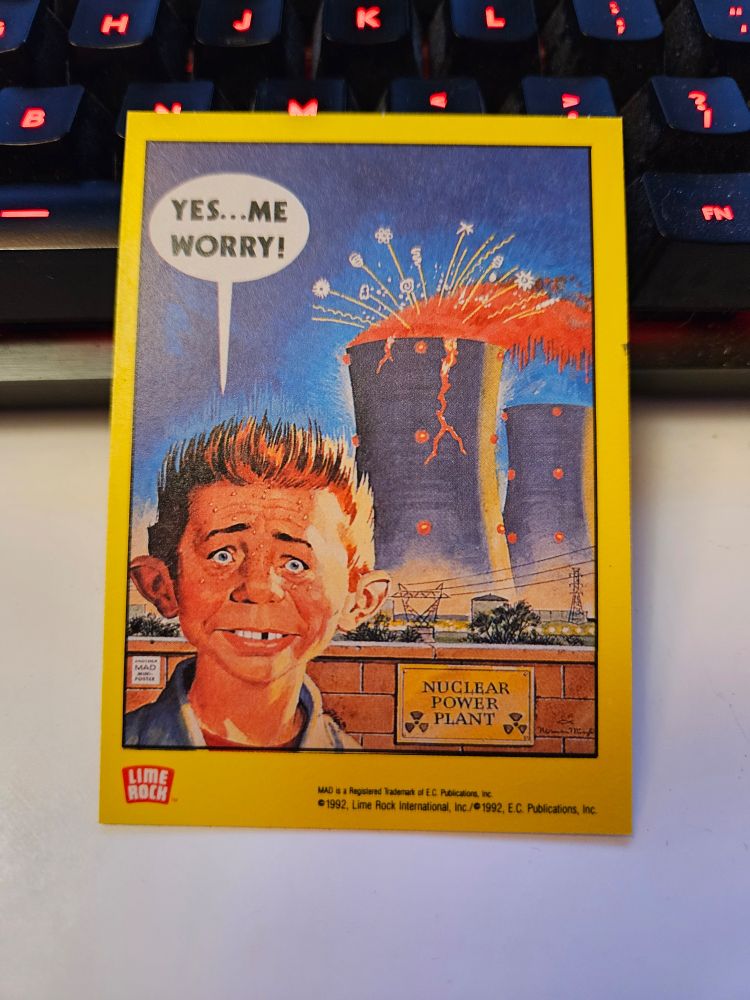 Mad magazine trading card showing Alfred E Newman in front of a nuclear power plant cooling tower that's on fire and cracking with a speech bubble above him reading "YES...ME WORRY!"