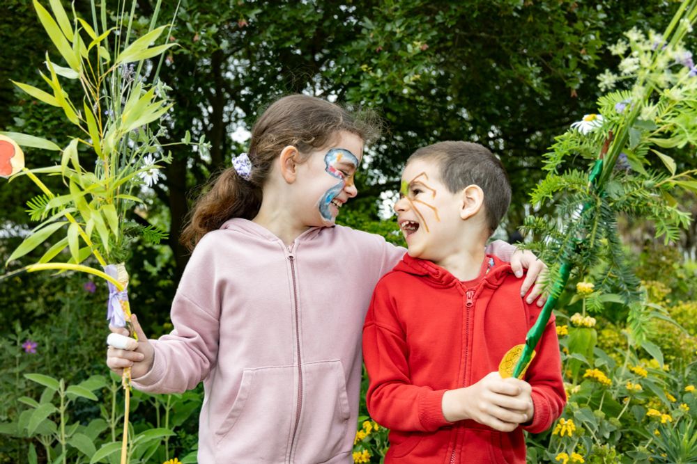 Two children looking at each other and laughing while waving plant wands that they have made at Cambridge University Botanic Garden