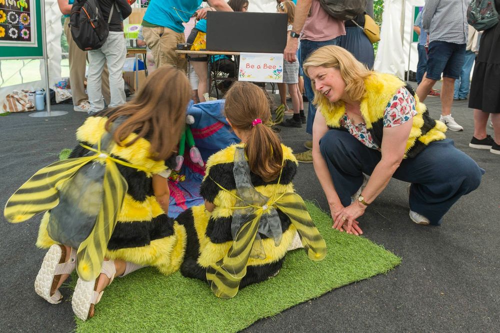 Two young girls dressed in bee costumes look at a model flower, whilst a smiling woman in a bee costume crouches near them.