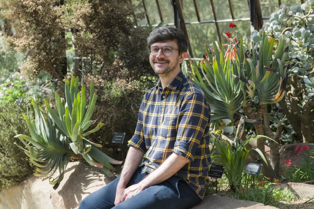 Matthew, in a blue and yellow plaid shirt, smiles as he sits in the Glasshouse Range at Cambridge University Botanic Garden