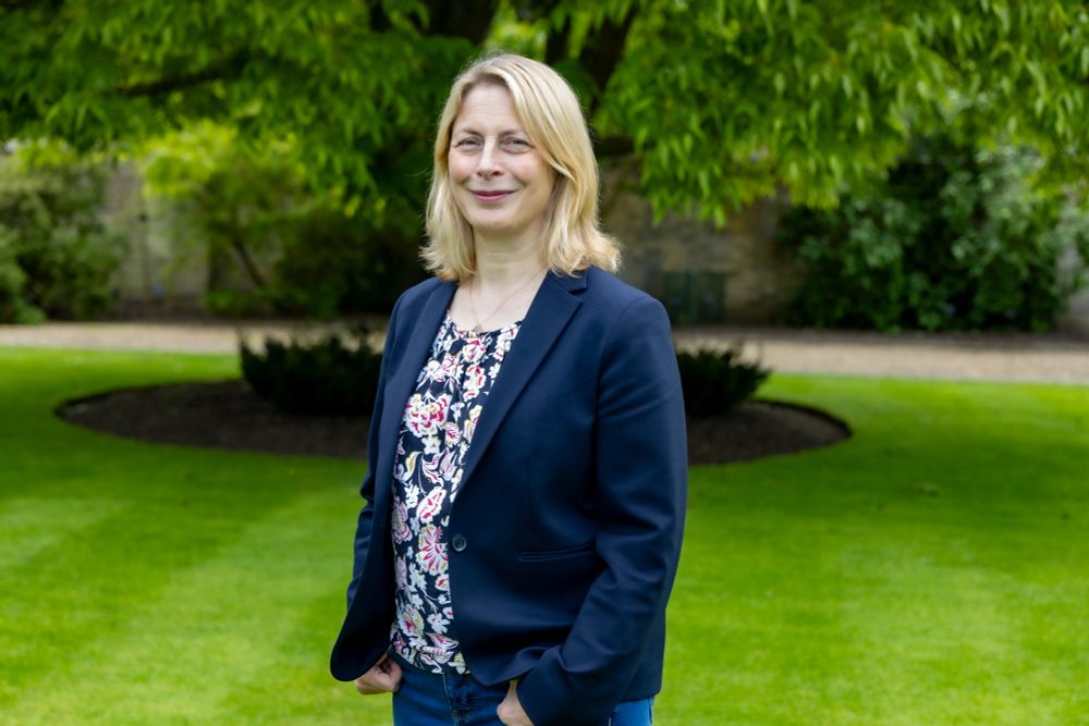 A woman in a blue blazer and floral top smiles whilst standing in a Garden.