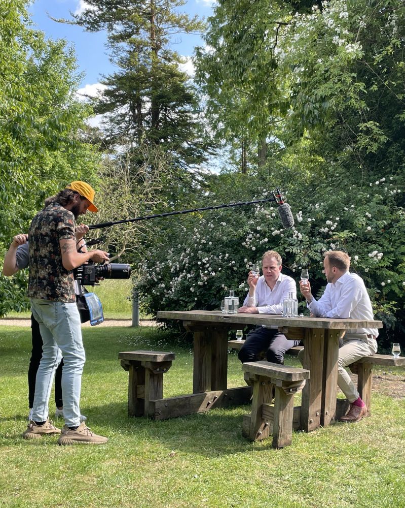 CUBG Curator, Sam Brockington and Cambridge Distillery Co-Founder William Lowe being filmed sitting at a picnic in CUBG tasting gin for the Drink documentary. A camera man and sound man can be seen to one side