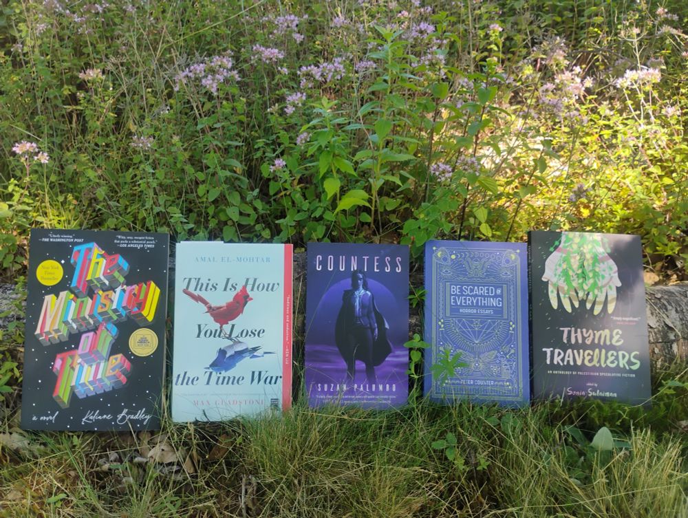A row of books lined up in front of sun-dappled plants with small purple flowers: The Ministry of Time, This is How You Lose the Time War, Countess, Be Scared of Everything, and Thyme Travellers.