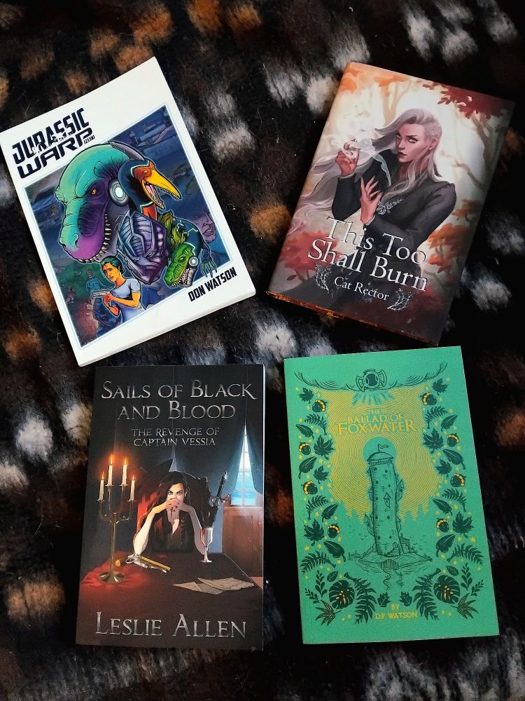 4 books over a brown bedding. "Jurassic Warp" is a comic book and "The Ballad of Foxwater" is a novel, both by Don Watson. "Sails of Black and Blood" is a novel by Leslie Allen and "This Too Shall Burn" is a novel by Cat Rector.