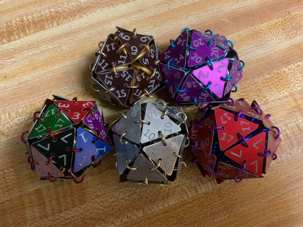 5 chainmaille d20s. Two on top are brown with copper rings and purple with blue rings. Bottom row is a mixed color of faces with red rings, silver faces with light yellow rings and red faces with purple rings.