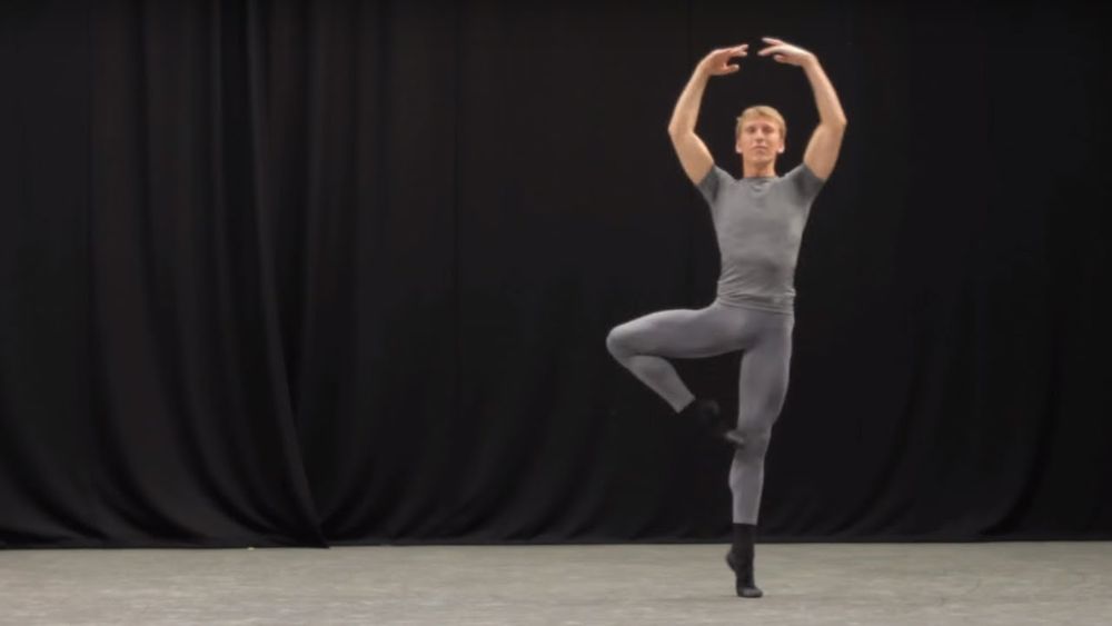 A male dancer in grey doing a pirouette against a black backdrop