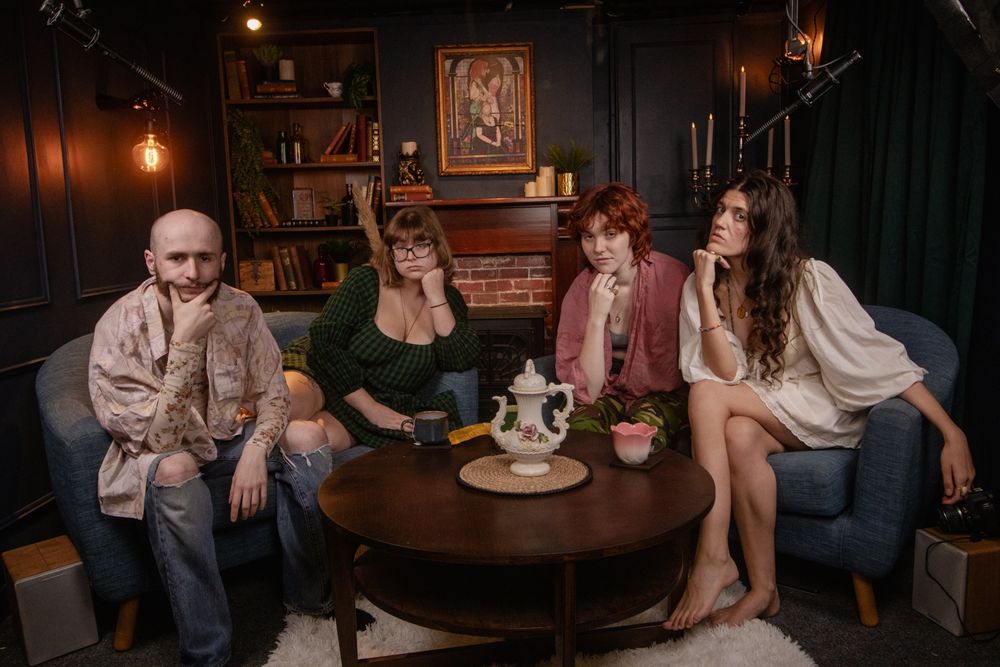 The Damsels recording studio lit by soft golden hanging lamps, with Rio, Morne, Maijai and Taya sat around on comfy blue sofas around a wooden table with the ornate white Teapot on it, all posing with hands under their chins as they stare intruigingly into the camera