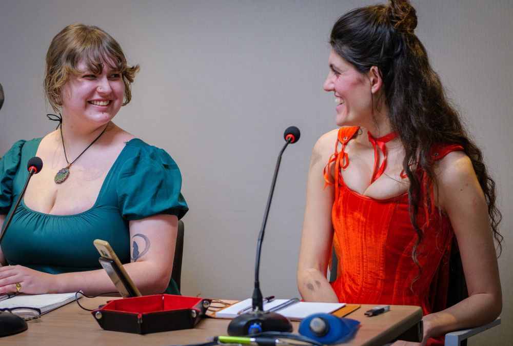 Morne on the right, in a green gown and necklace with their blonde hair tied back, laughs with Taya on the left, in a bright red corset and ribbon, long dark hair half up half down, covered in rogue and dirt. They sit at a table with mics and dicetrays.