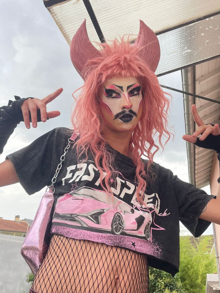 Artiste drag (moi) avec une perruque rose à cornes, un maquillage drag inspiré goth avec des nuances de rose, une moustache, un crop top noir avec une voiture rose dessus, un sac a main rose métallique, des résilles couvrant son ventre, et faisant des gang signs pointant vers son visage.