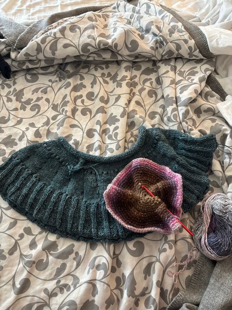 A topper handknit in 2 colors of Sea Change Shoreline Sport (Aquatic and Sea Hag, both very close blue-greens) sits on a bed; a crocheted hat underway in Noro Silk Garden colorway unknown in shades of brown, gray, purple and pink. 