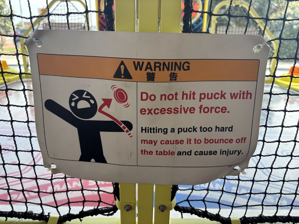 A warning sign on a Pac-Man air hockey machine. It reads "Do not hit pick with excessive force". A picture shows a stick figure crying as a puck hits him with excessive force.