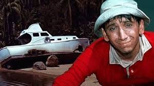 Gilligan (Bob Denver)  in red shirt over white shirt and white bucket hat sitting at the beach with the shipwrecked white "SS Minnow"  in the background