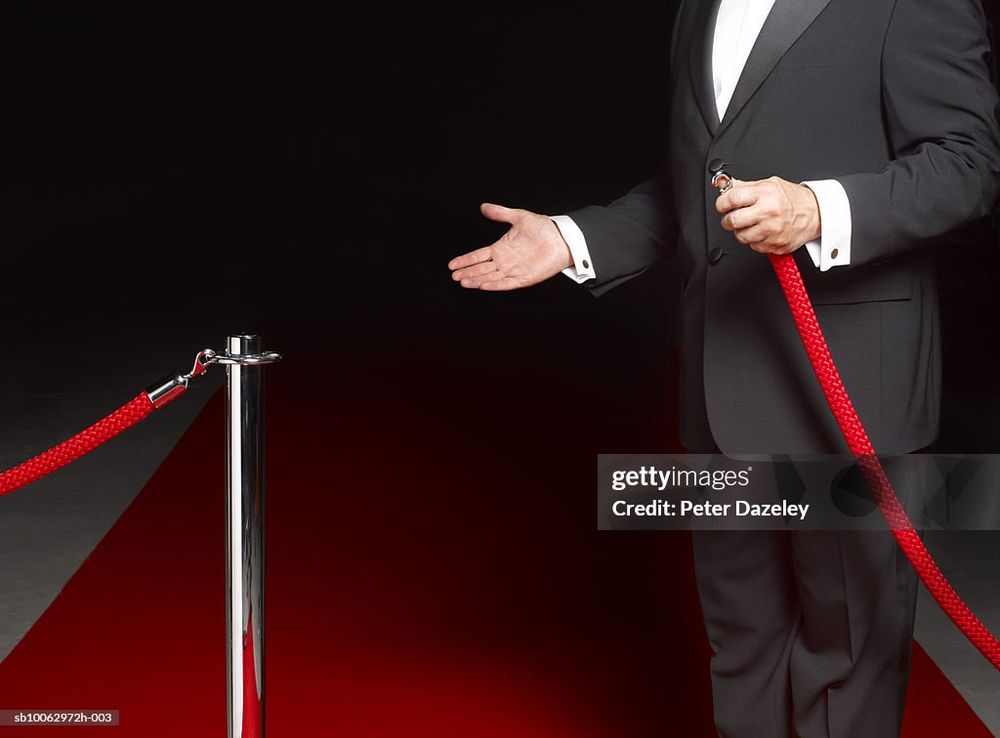 a man wearing a black tuxedo readying to hook red velvet rope to stanchion at left of screen