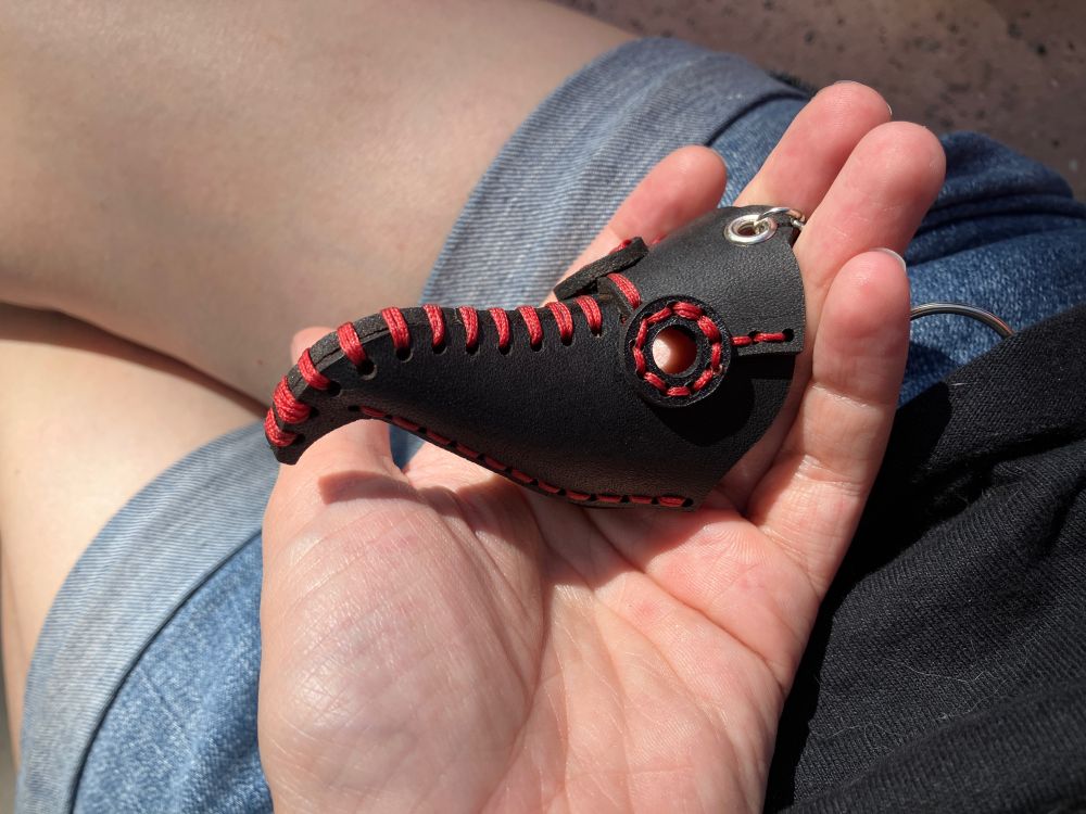 A little black leather plague doctor mask keychain with red stitching is being cradled in a loving palm