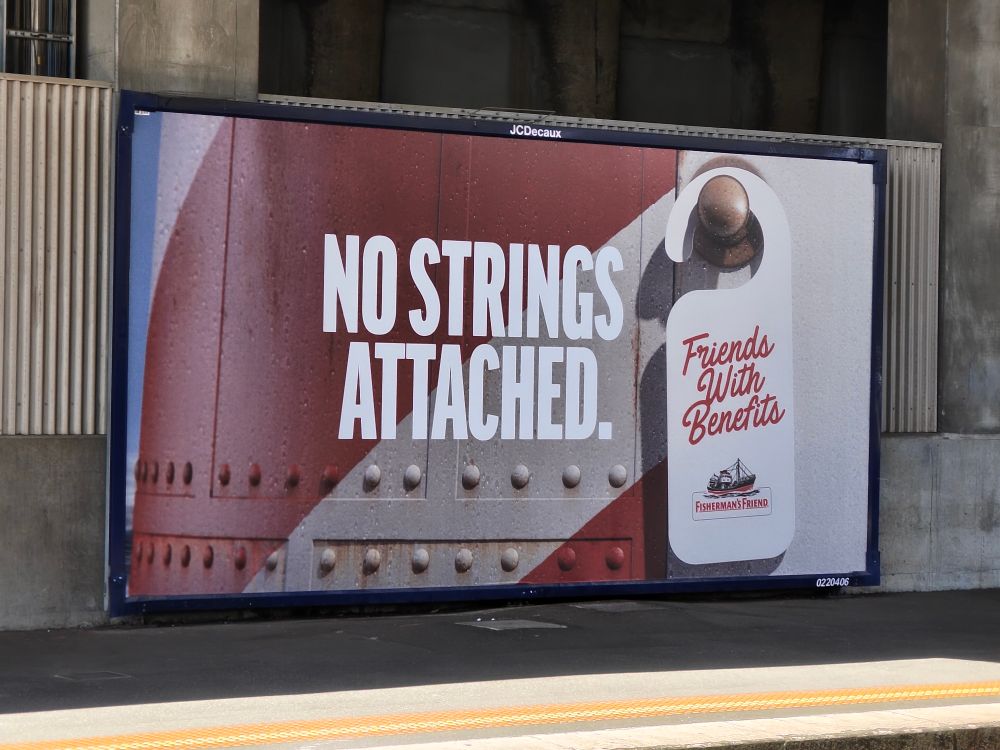 billboard ad at a train station for fisherman's friend. it's a light house with a door hanger saying "friends with benefits" and the caption next to it "no strings attached"
