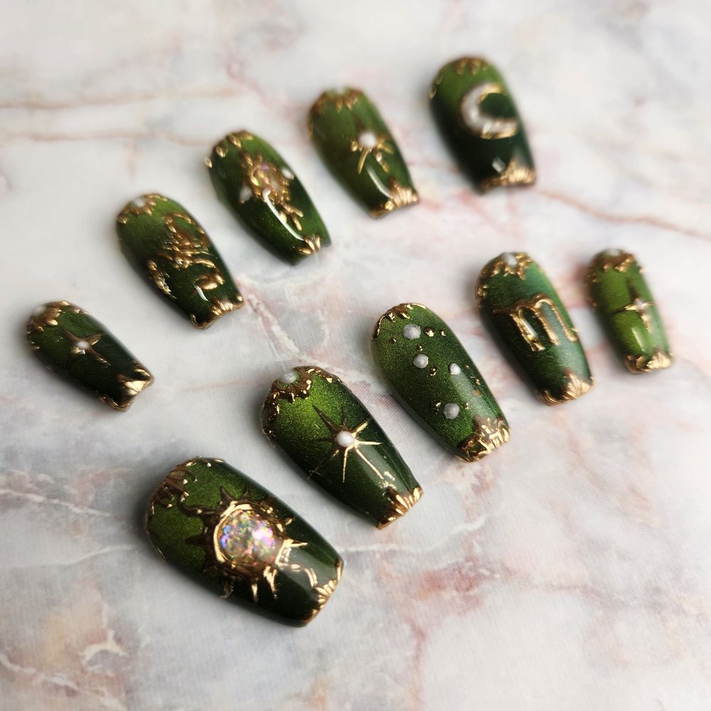 Set of short, coffin shaped press-on nails on a light marble background. The nails have an olive green shimmery base, with gold celestial and Scorpio themed designs.
