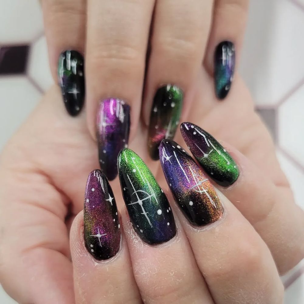 Close of two hands, displaying nail art. Nails are in a rounded stiletto shape, with a black base, with multi-colored nebulas, and white stars.