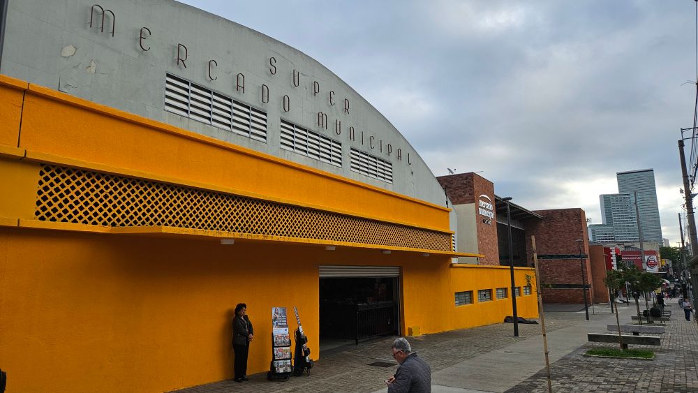 Foto da fachada do mercado municipal da cidade..eh uma construção amarela, seguida de um outro prédio (provavelmente a expansão)