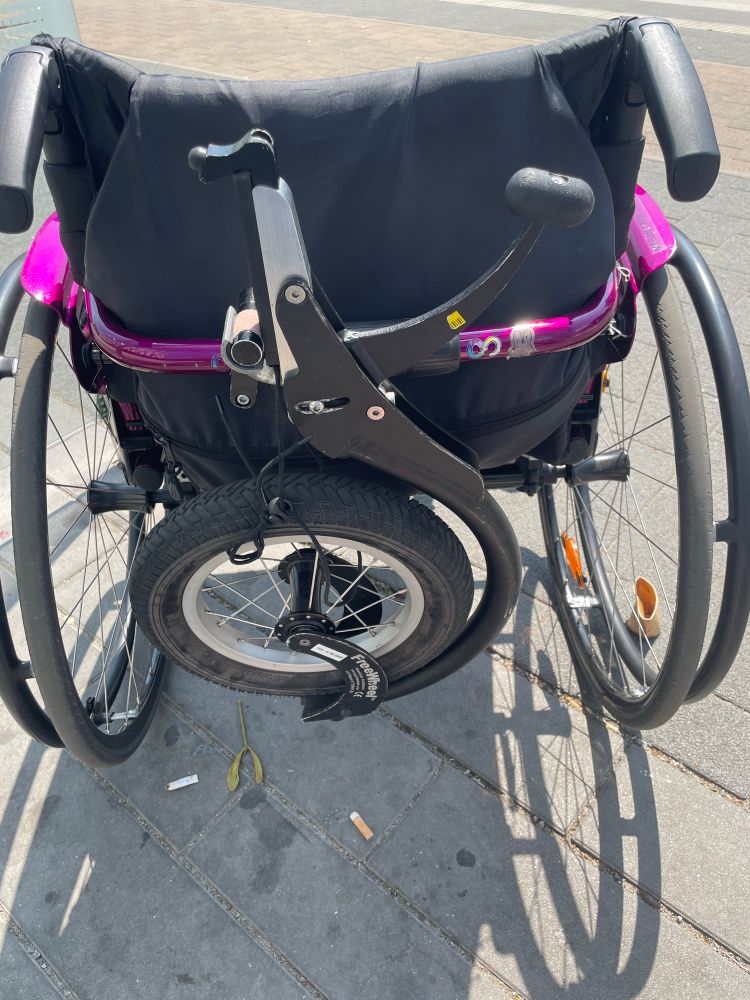 Rear view of a wheelchair with an extra all terrain wheel clipped on the back. 