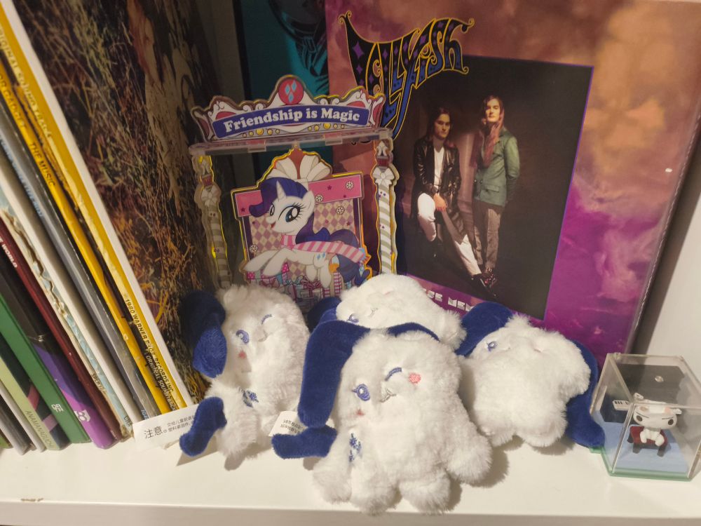 a photo showing an acrylic stand of rarity from my little pony g4, and four plush toys of the same character. 
(unrelated to the subject of this post: a figure of toro inoue, the boxset of 7" jellyfish singles "when these memories fade", the 12" "scary-go-round ep" also by jellyfish, a collector's edition of gorillaz's "the now now", and the spines of various other records.)