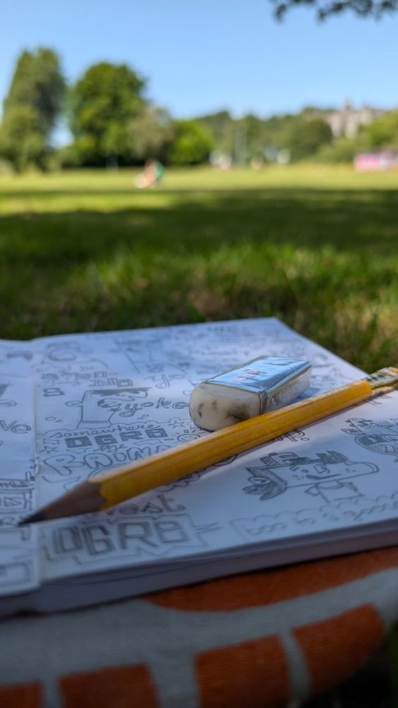 A sketch book and pencil on grass, with green parkland and blue sky in the background 