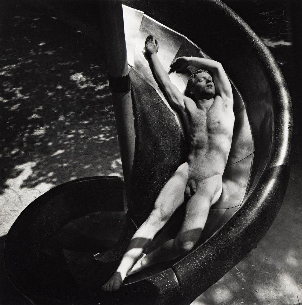 A nude man lies stretched out on a curving playground slide, with dappled sunlight casting shadows on his body.