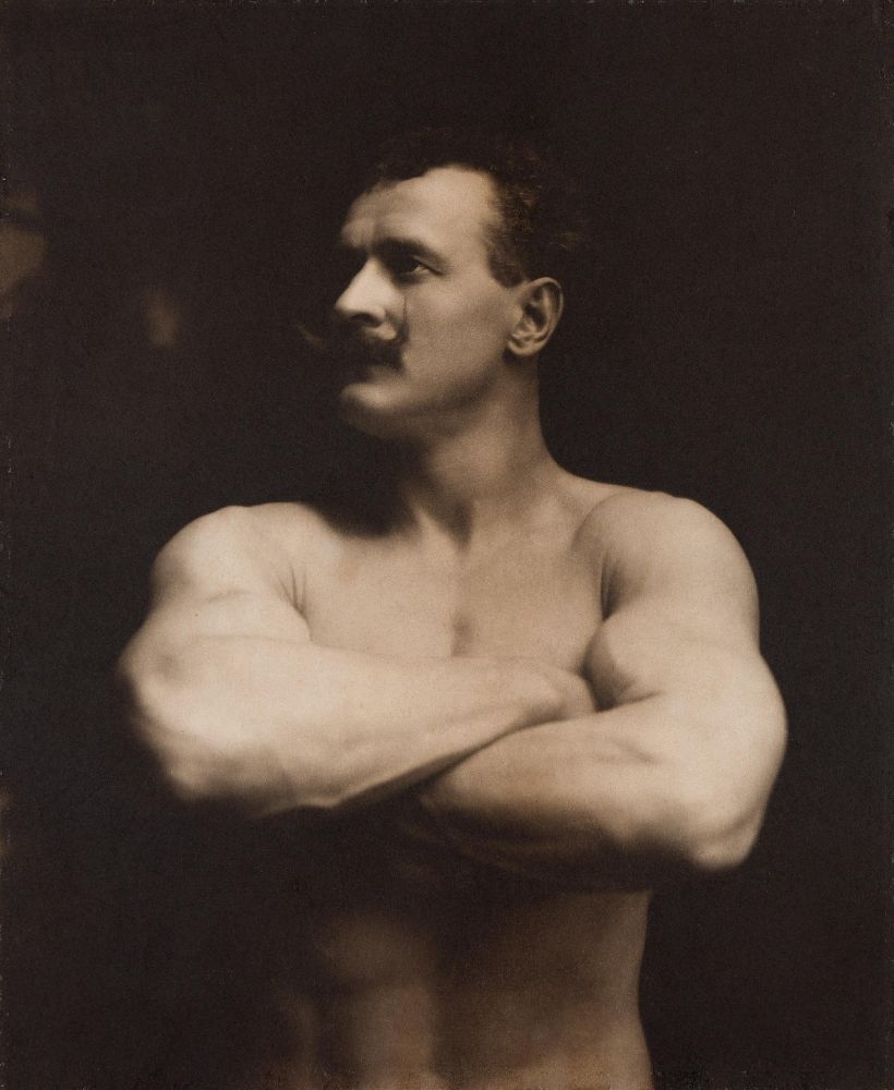 Shirtless man with a muscular build and a prominent mustache poses with arms crossed, gazing to the side in a sepia-toned portrait.