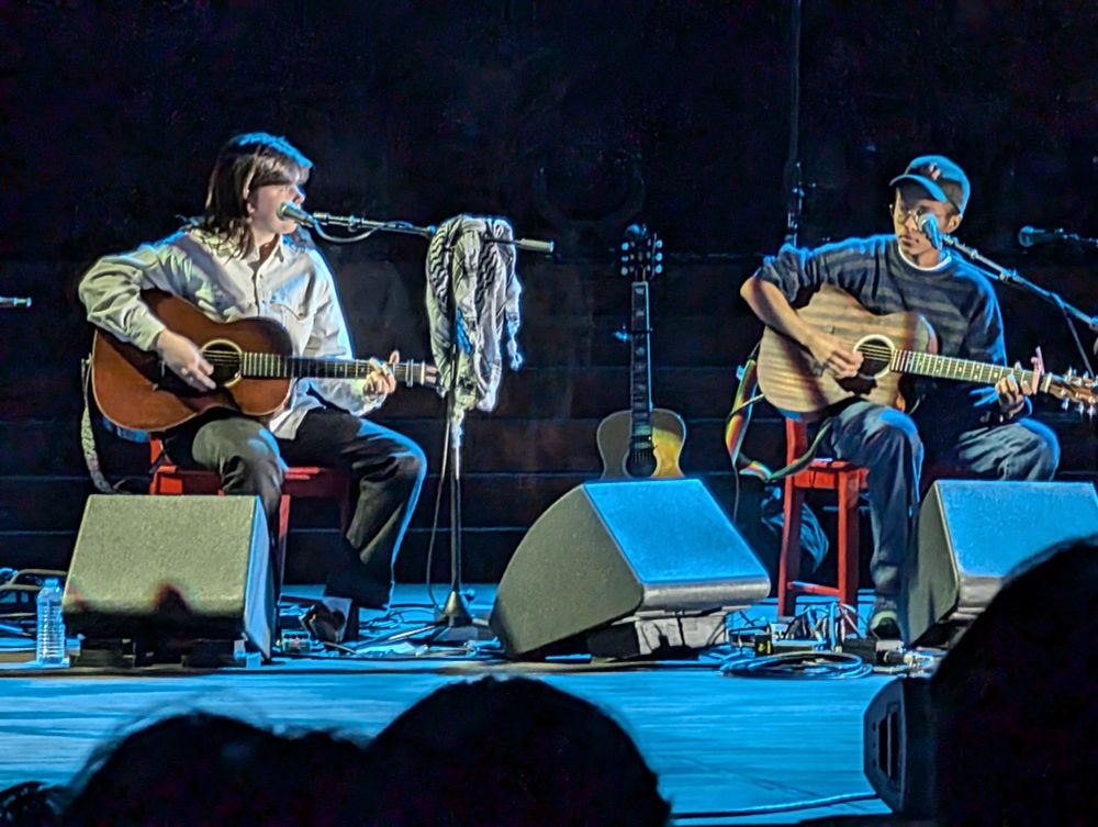 Leith Ross holding a guitar in front of a microphone with a keffiyeh. Rae Chen, also known as the artist Tofusmell, sits next to them. Rae also holds a guitar.