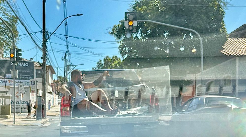 Foto de um homem sentado na caçamba de uma caminhonete, com a porta aberta, segurando uma porta de vidro solta, sem nenhum equipamento de proteção.