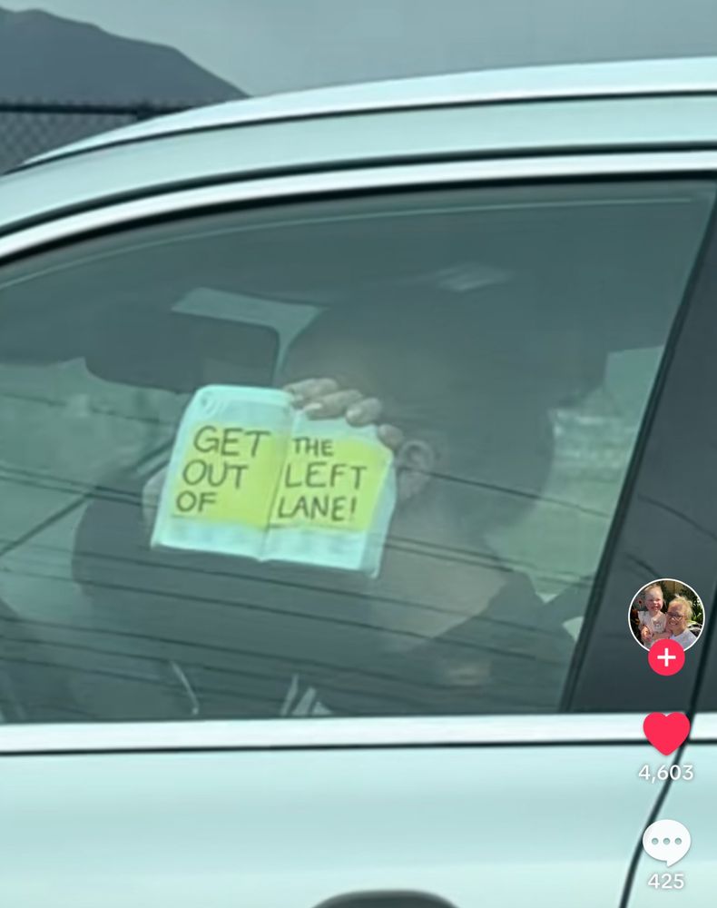 Woman, while driving on I-15 in what appears to be Weber County, Utah is holding up an open copy of the Book of Mormon to her left window, so the occupants in the vehicle in the left lane next to her can see. On two yellow “stickies” in the open book, are written “GET OUT OF THE LEFT LANE”